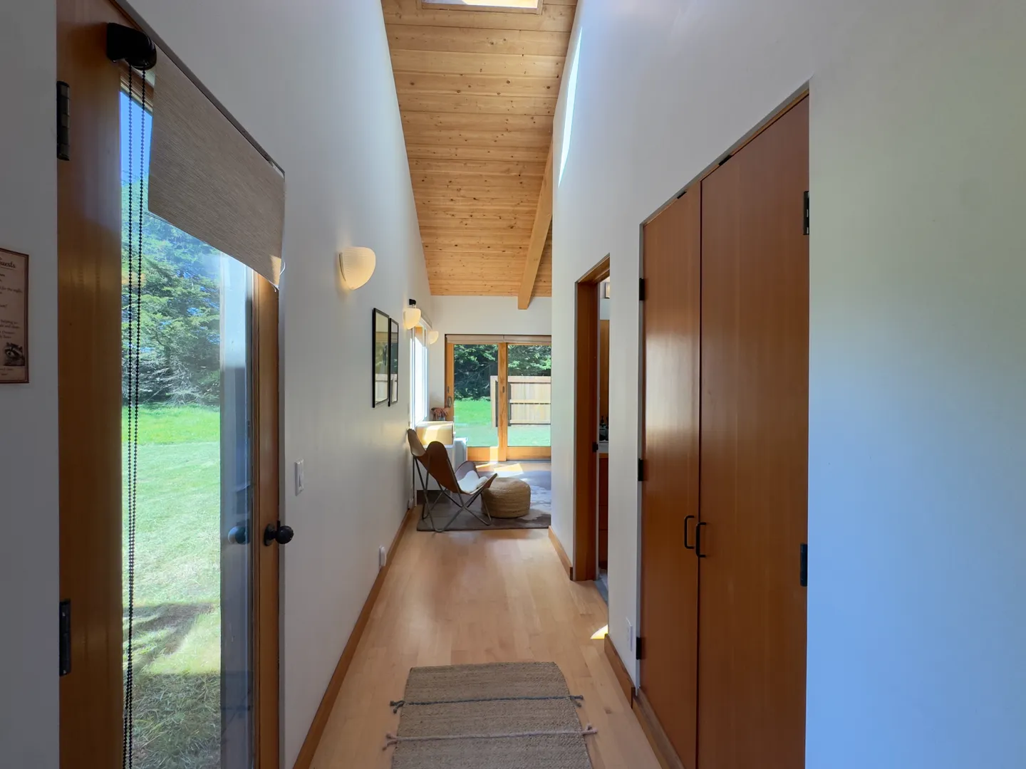 Hallway with light wood flooring, vaulted wood-paneled ceiling, white walls, and glass doors looking out to a green yard.