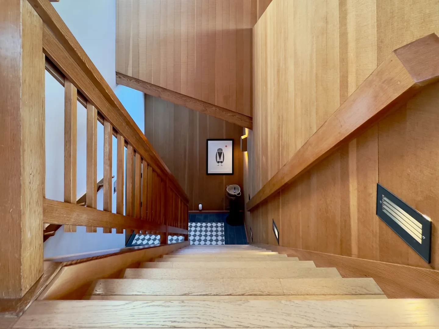 Looking down a wooden staircase flanked by wood-paneled walls and a wooden railing toward a lower landing with patterned tile.