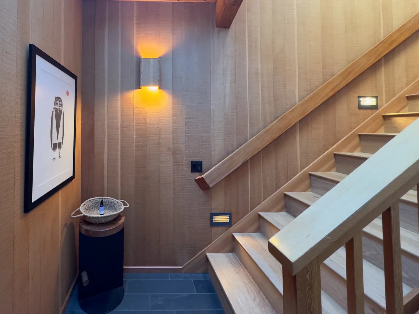 A wooden staircase with wood-paneled walls, warm sconce lighting, and framed artwork on the landing.