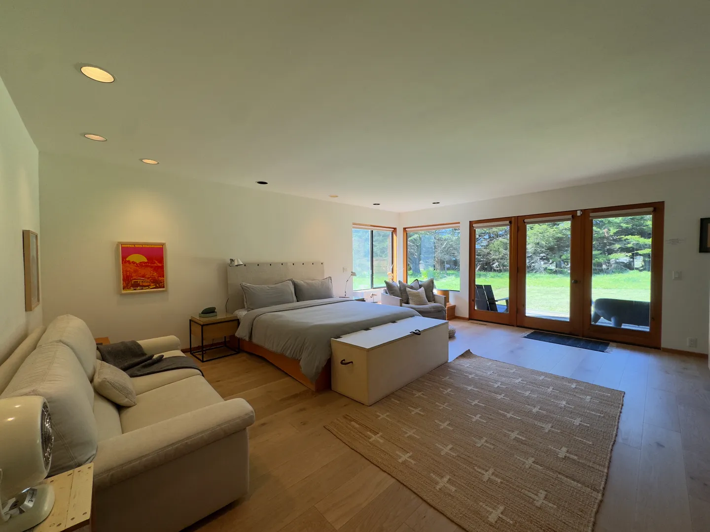 Spacious bedroom with wood flooring, a bed, a sofa, an armchair, and glass doors opening to a grassy yard.
