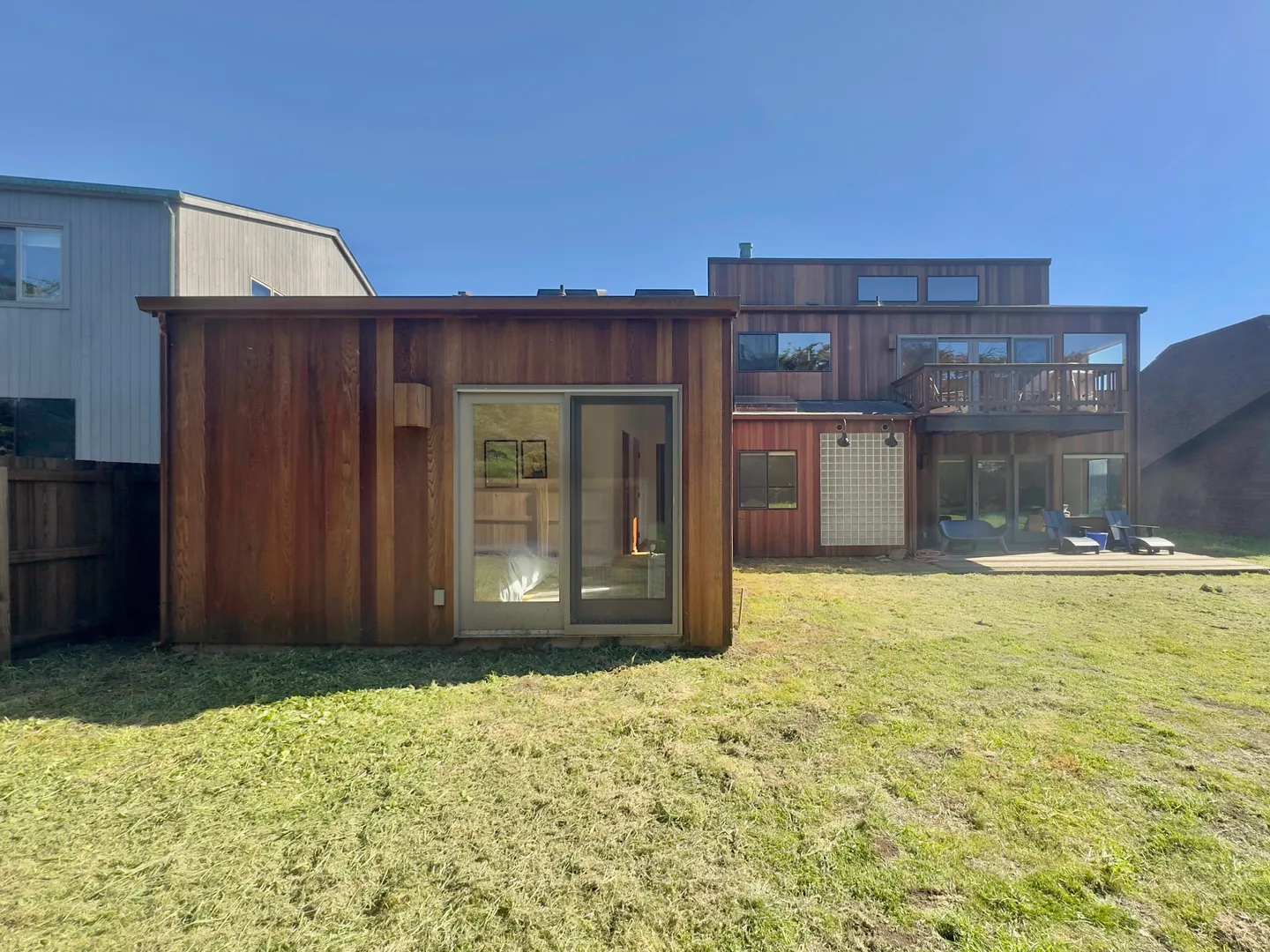 Rear exterior view of a multi-story home with natural wood siding, an open grassy yard, and an attached deck with lounge chairs.