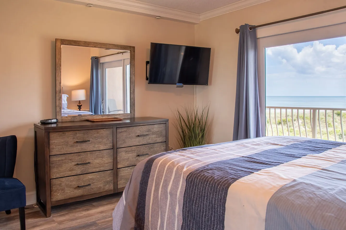 Bedroom with a bed in blue striped bedding, a wooden dresser with a mirror, a wall-mounted TV, and sliding glass doors revealing an ocean view.