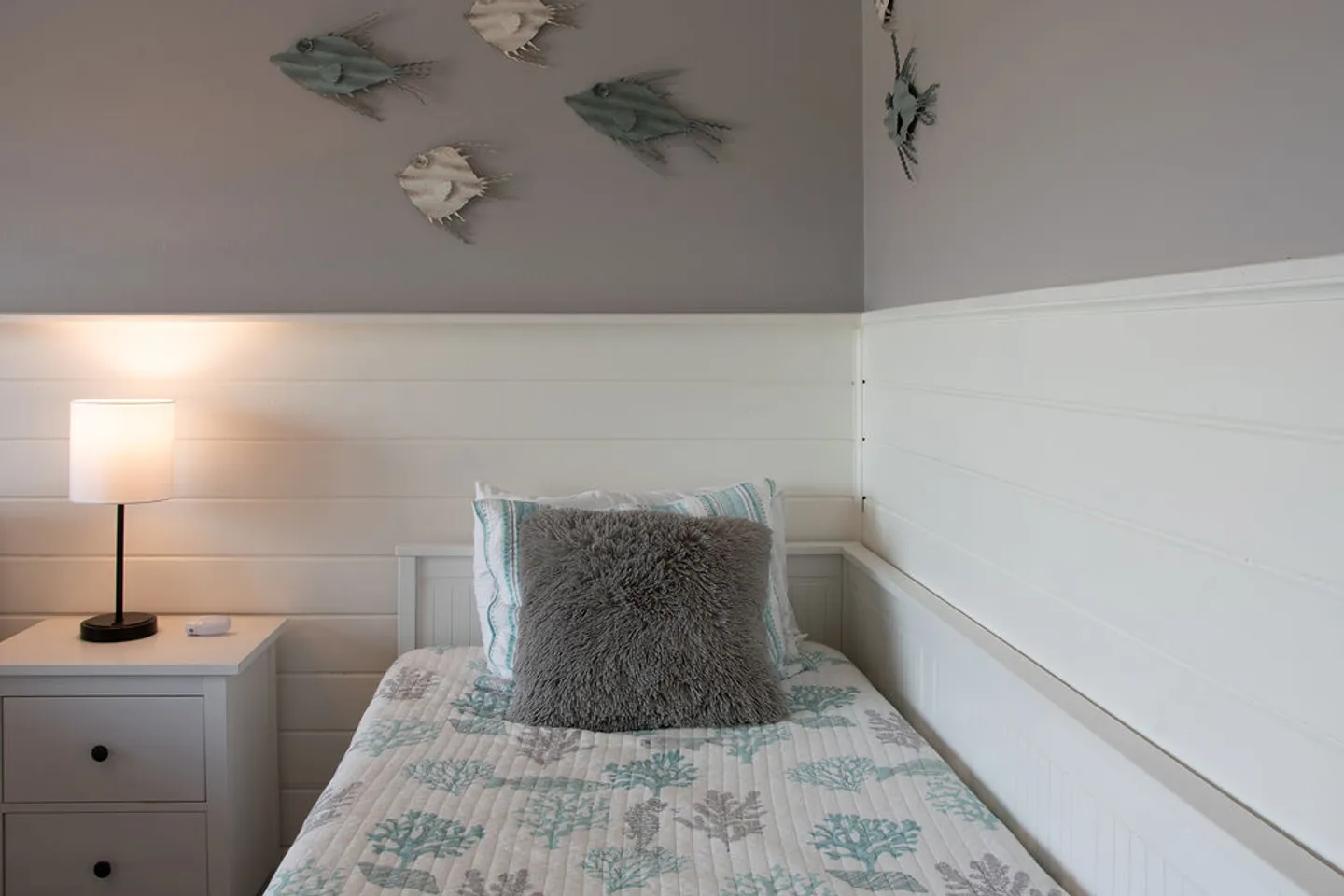 A corner bed with coastal-themed bedding, white shiplap wainscoting, and decorative fish mounted on the wall above.