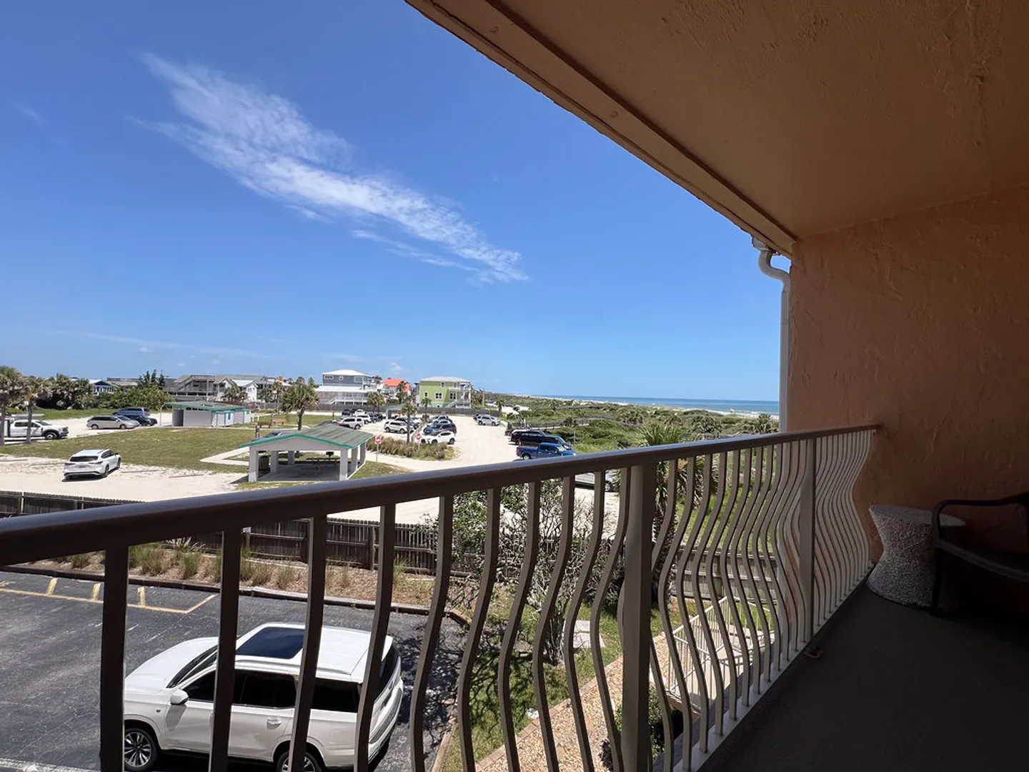 A covered balcony with curved white railings looks out over a parking lot and coastal dunes toward the ocean under a blue sky.