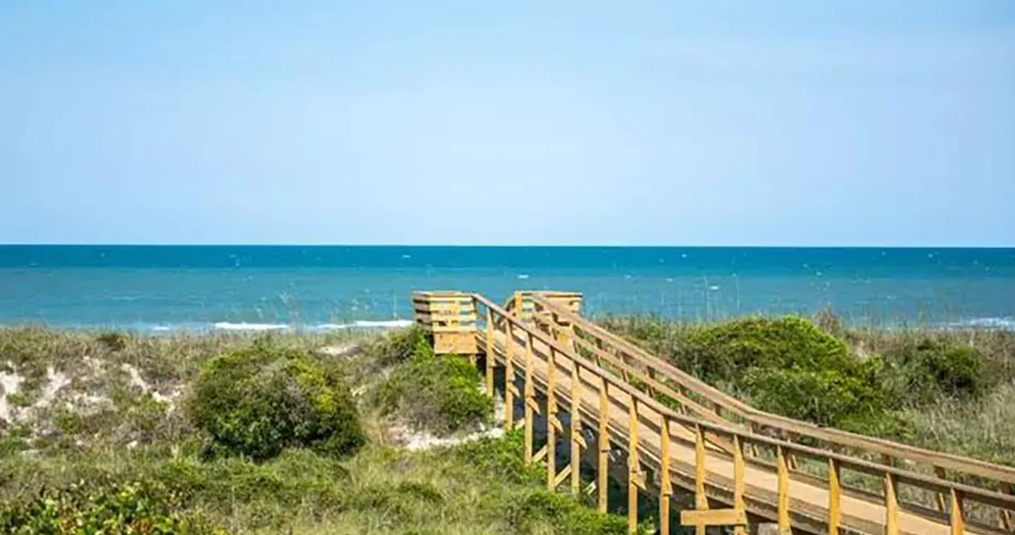 Wooden boardwalk with railings leading across grassy sand dunes toward the ocean under a clear sky.