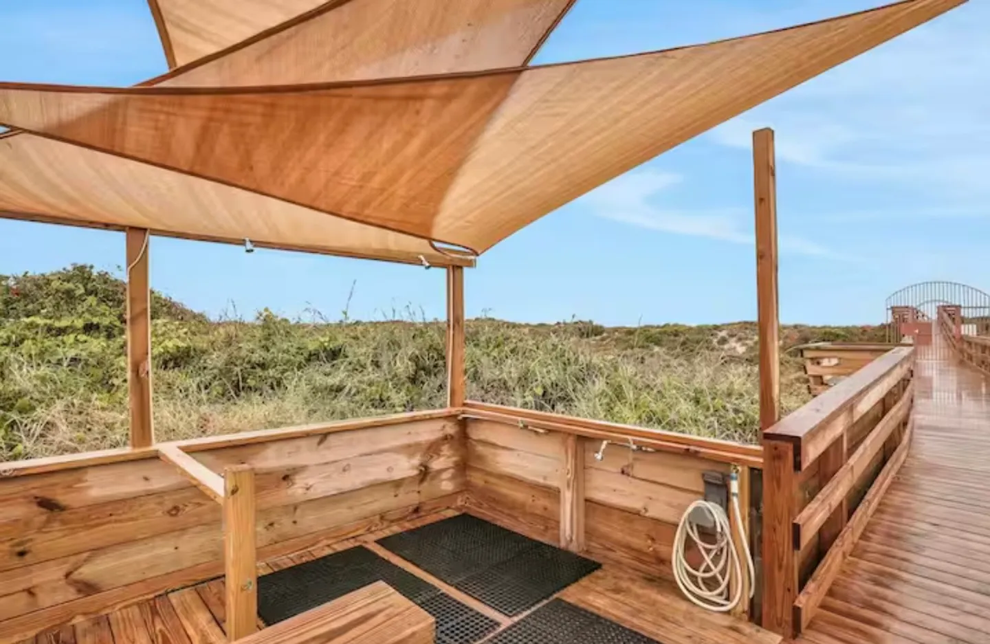 Outdoor wooden rinse-off station with showerheads and foot wash under shade sails, situated along a boardwalk.