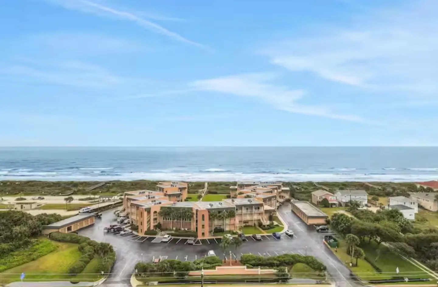 Aerial view of an oceanfront condominium complex with parking lot, grassy grounds, and direct beach access under a partly cloudy sky.