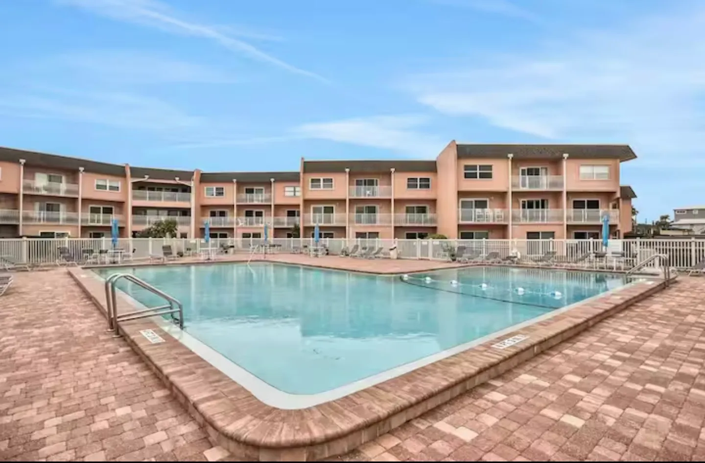 Large outdoor community swimming pool surrounded by brick pavers with multistory pink condominium buildings in the background.