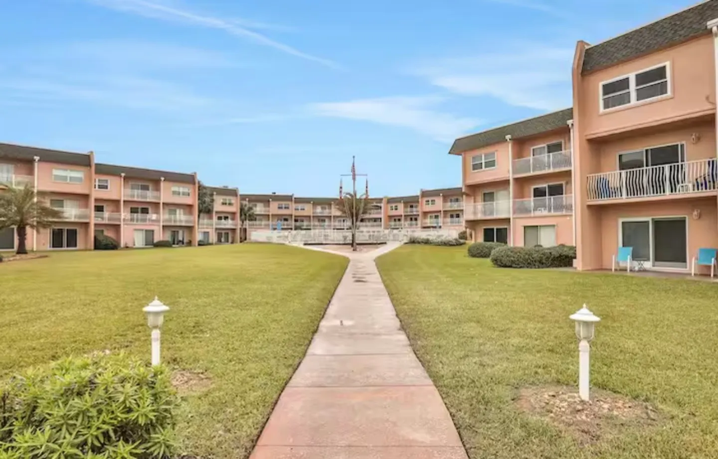 A concrete walkway leads through a central grass lawn surrounded by multi-story, peach-colored condominium buildings under a partly cloudy sky.