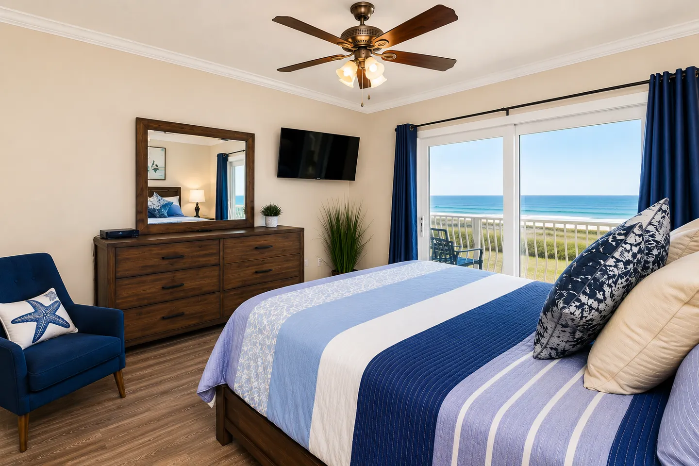 Bedroom with a bed made with blue bedding, a blue armchair, a dresser with mirror, and sliding glass doors opening to a balcony with ocean views.