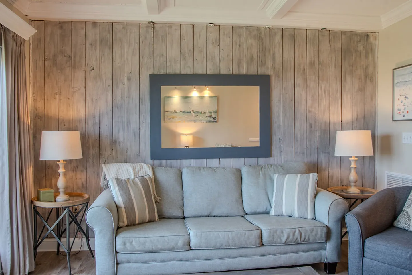 Living room seating area with an upholstered couch, accent pillows, end tables with lamps, and a wood-paneled accent wall with a mirror.