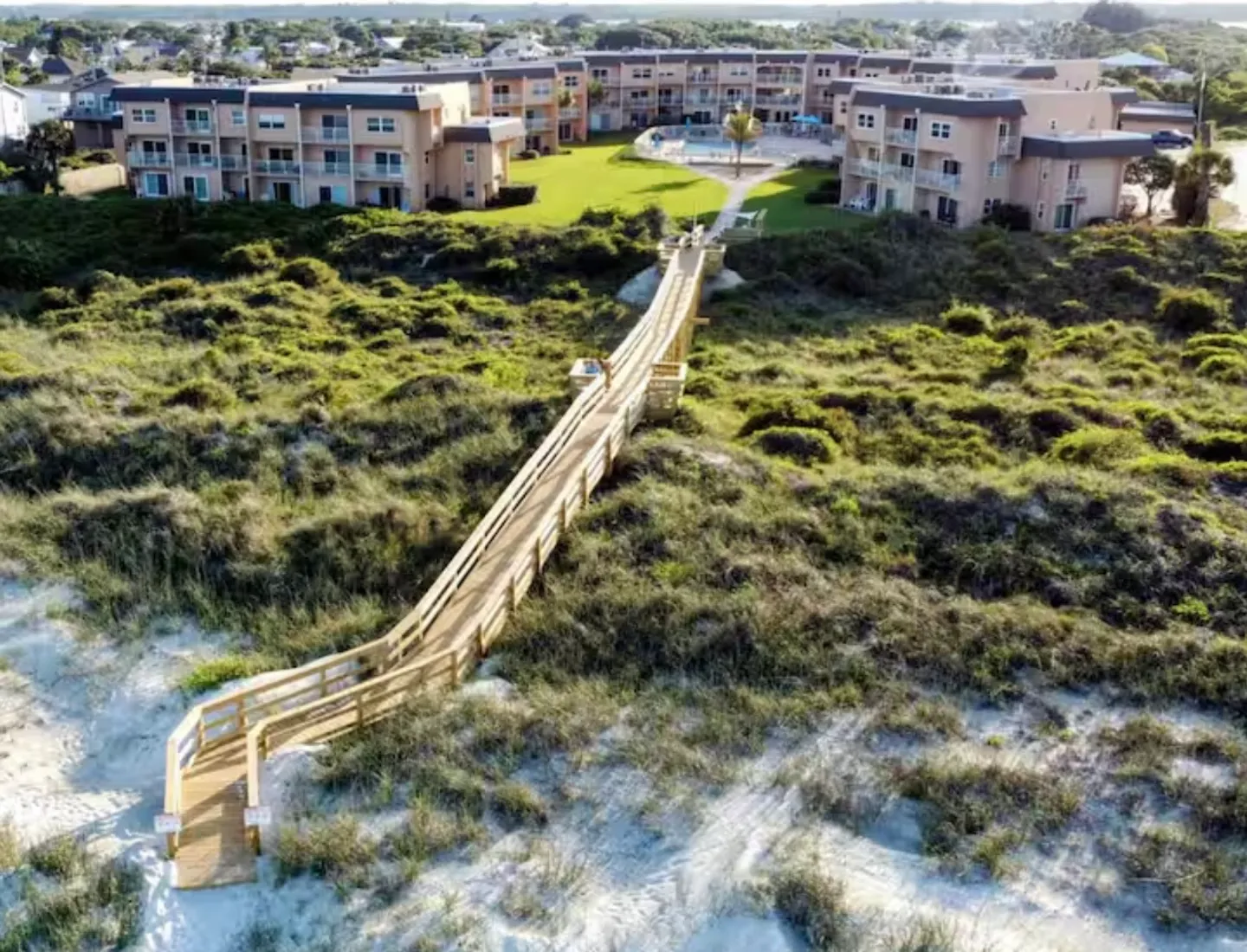 Aerial view of a long wooden boardwalk crossing dunes between sandy beach grass and a multi-building condominium complex with a pool.