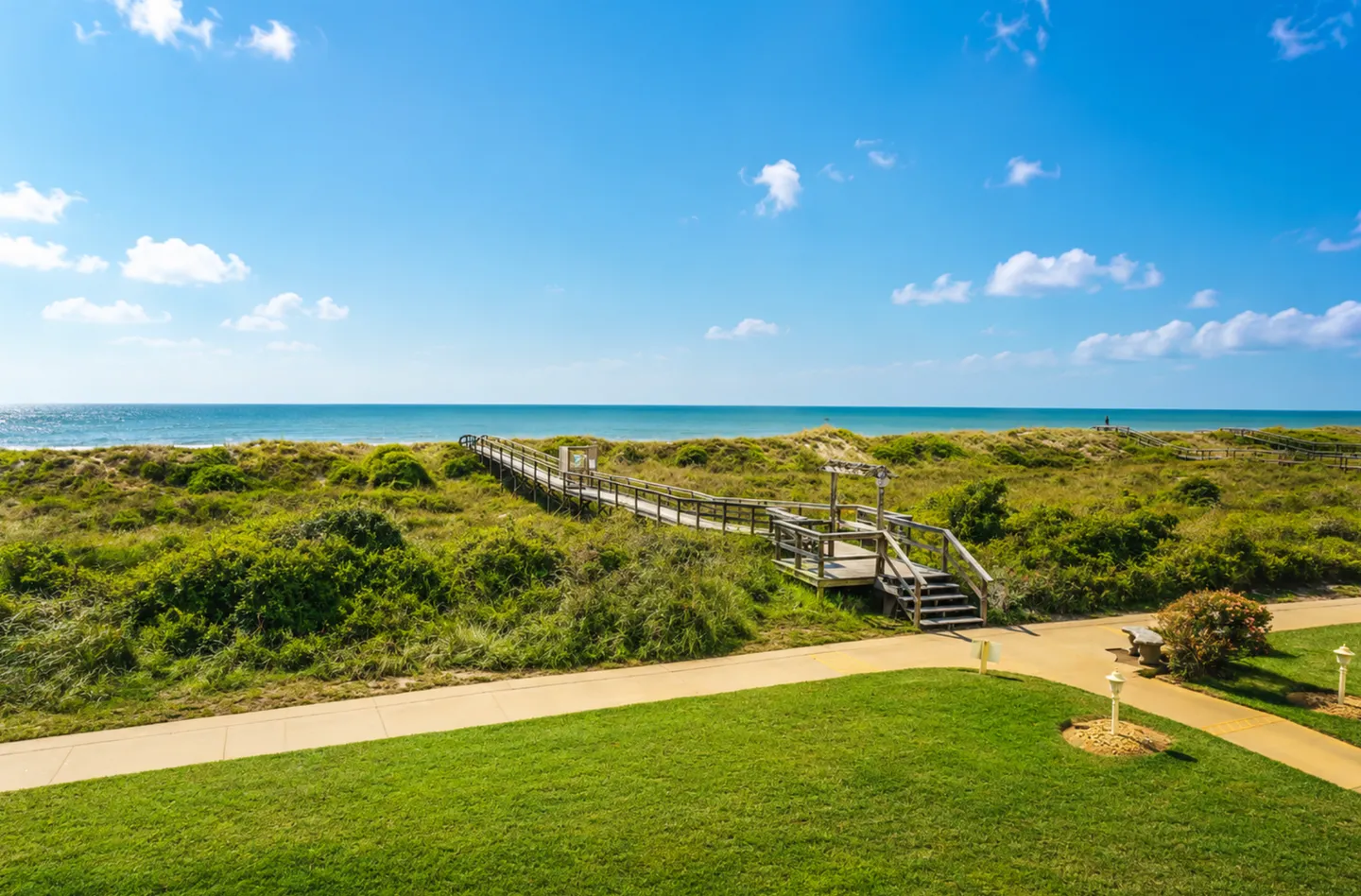 A wooden boardwalk leads through sand dunes and coastal vegetation toward the ocean beneath a clear blue sky.