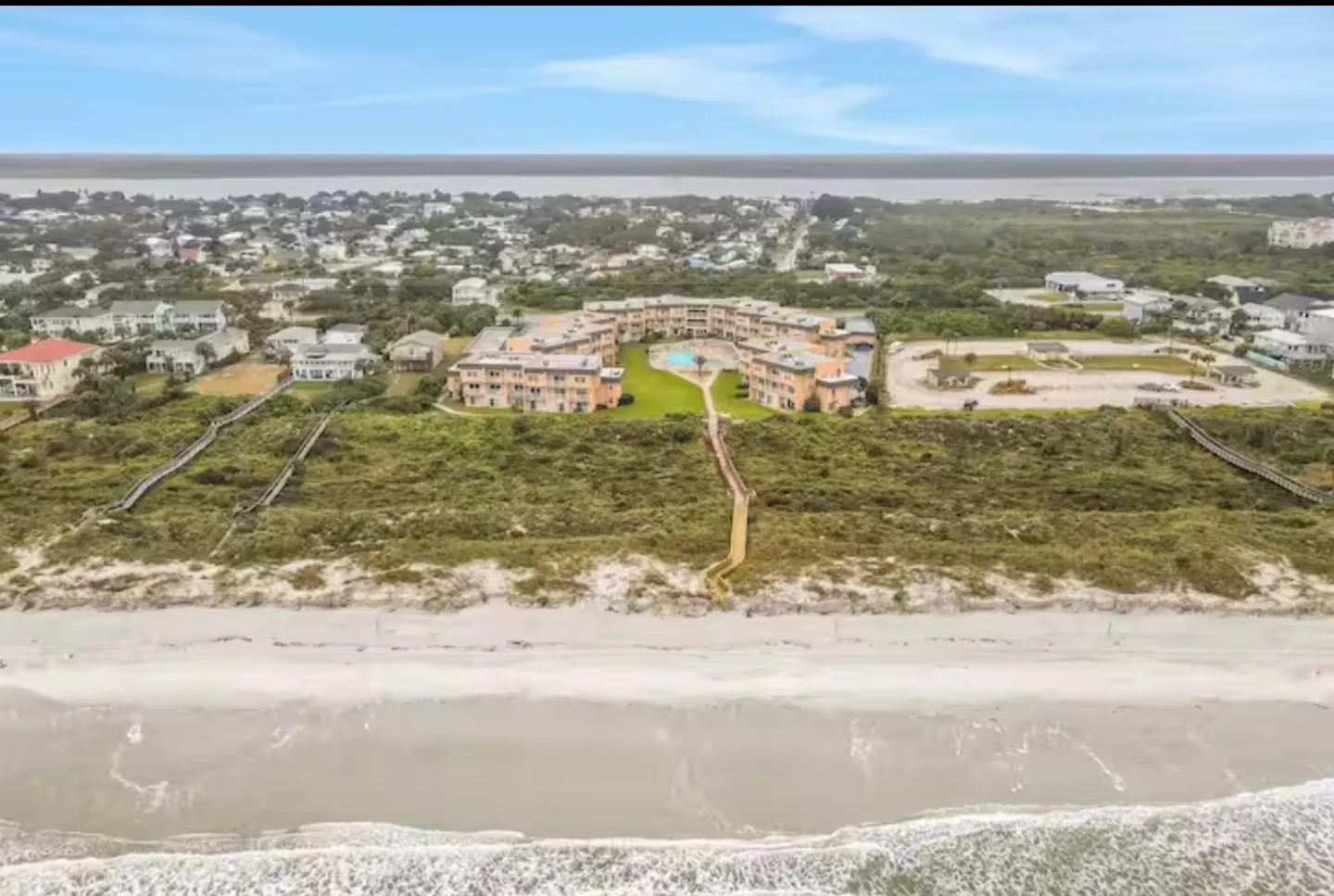Aerial view of an oceanfront condo complex featuring a central swimming pool and wooden boardwalk extending across the dunes to the sandy beach.