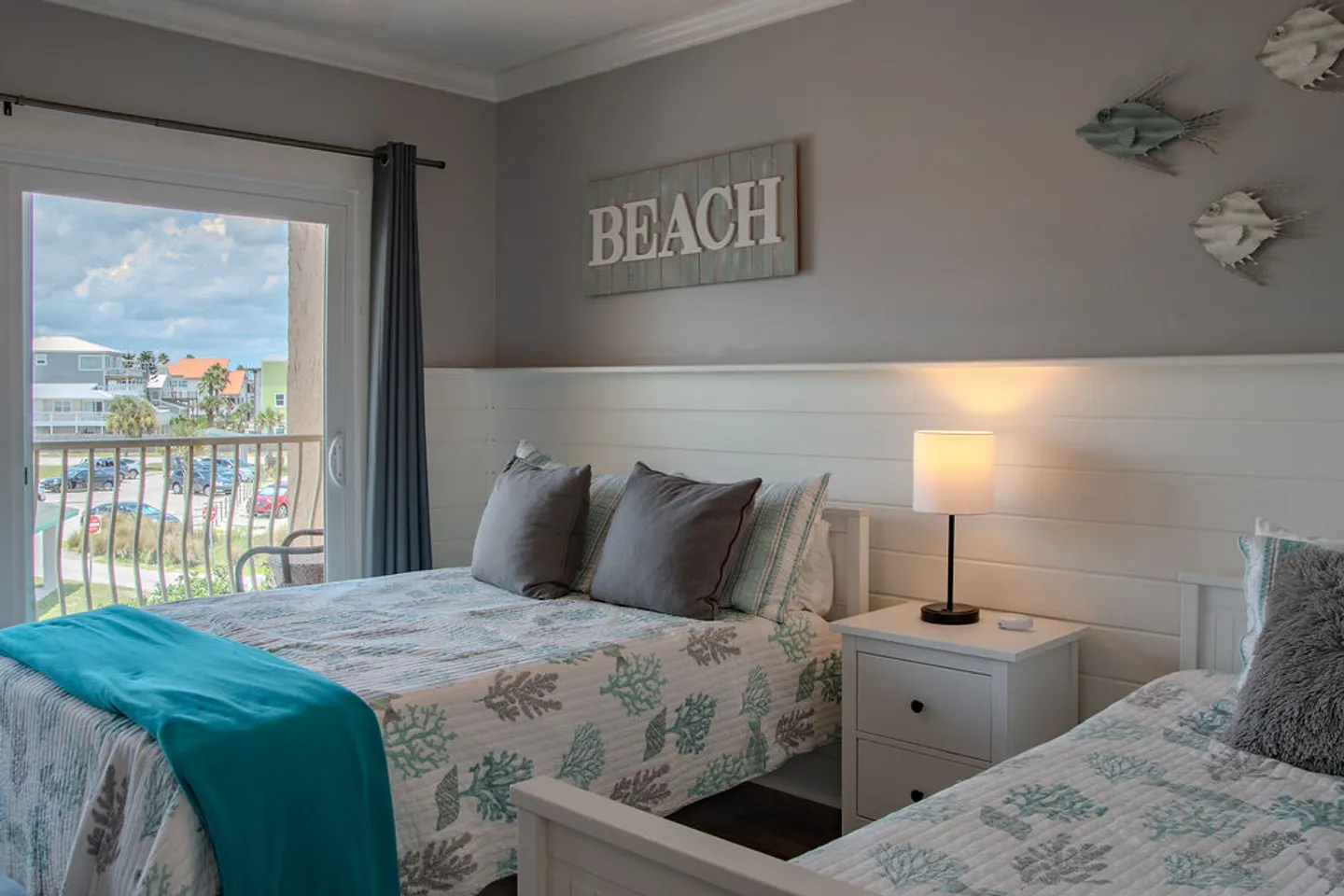 Bedroom with two beds, coastal-patterned bedding, a nightstand with a lamp, and a sliding glass door leading to a balcony.