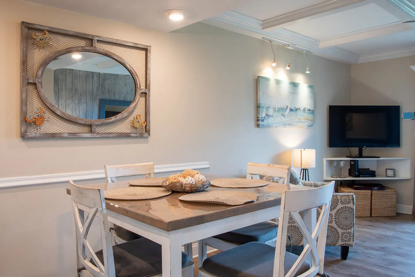 Dining area with a square wooden table, placemats, and four chairs, opening to a living space with a television.