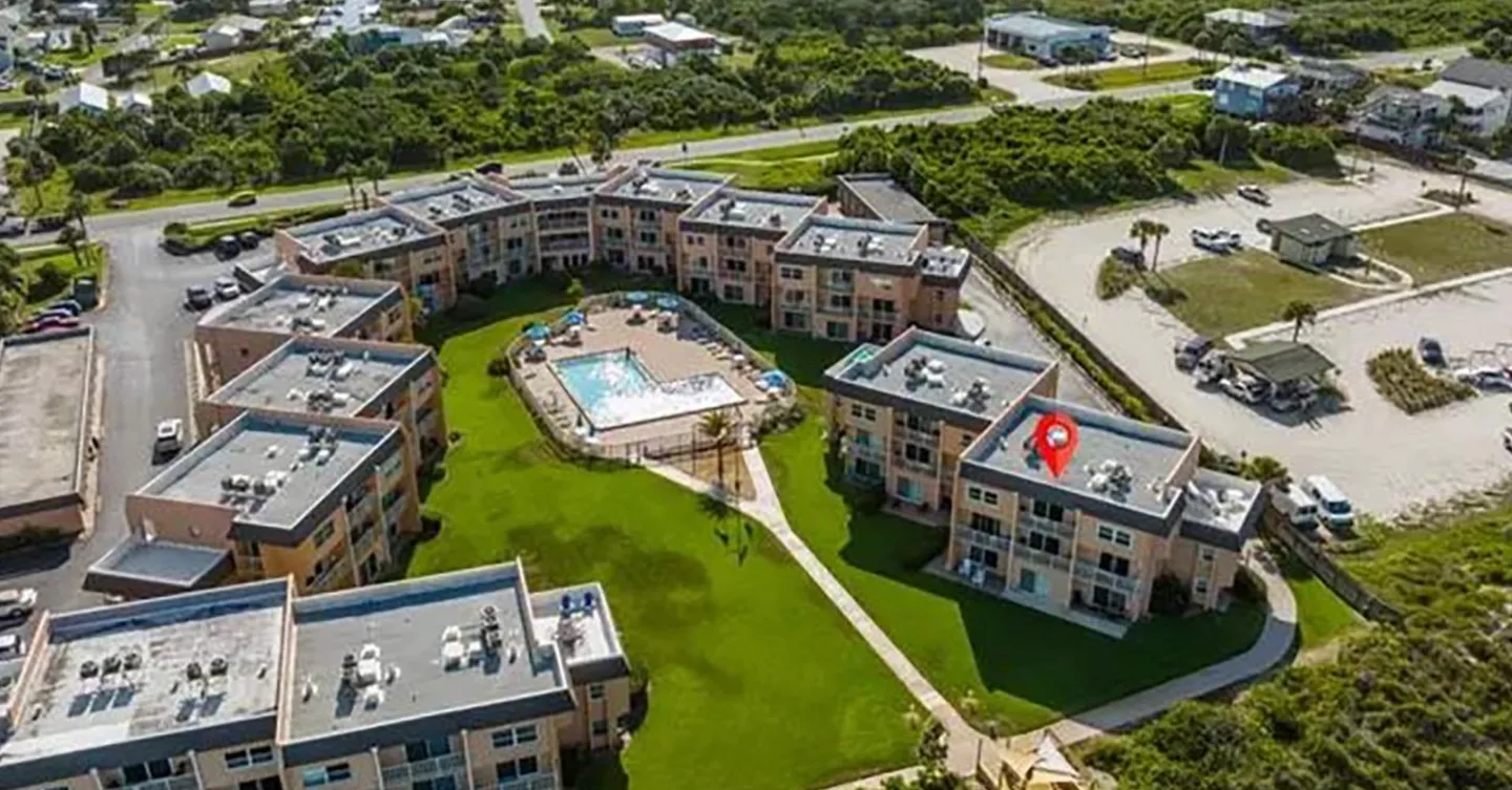 Aerial view of a condominium complex surrounding a central swimming pool and lawn, with a red pin on one building.