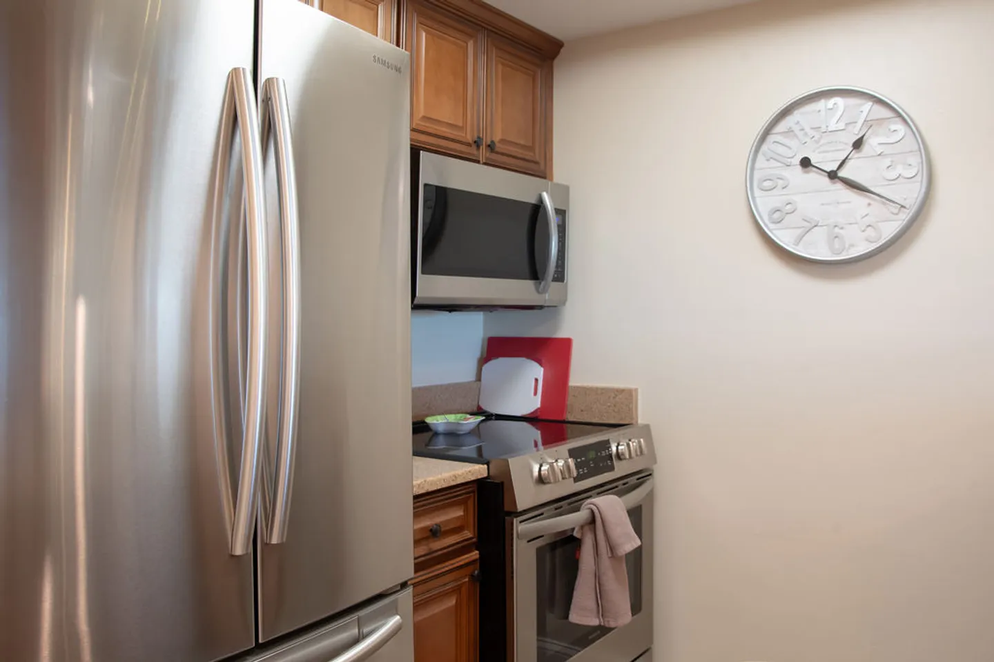 Close-up view of kitchen area featuring a stainless steel refrigerator, microwave, electric range, wood cabinetry, and a large round wall clock.
