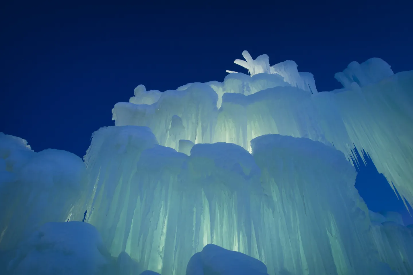 Low-angle view of a large ice castle structure illuminated from within against a dark blue evening sky.