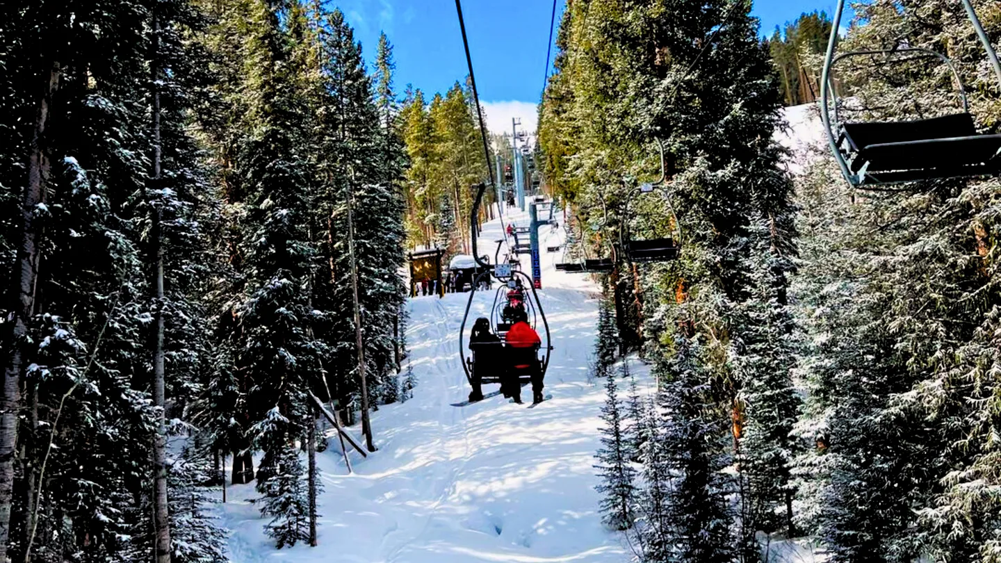 A view from a ski chairlift ascending a snow-covered mountain slope surrounded by tall pine trees under a clear blue sky.