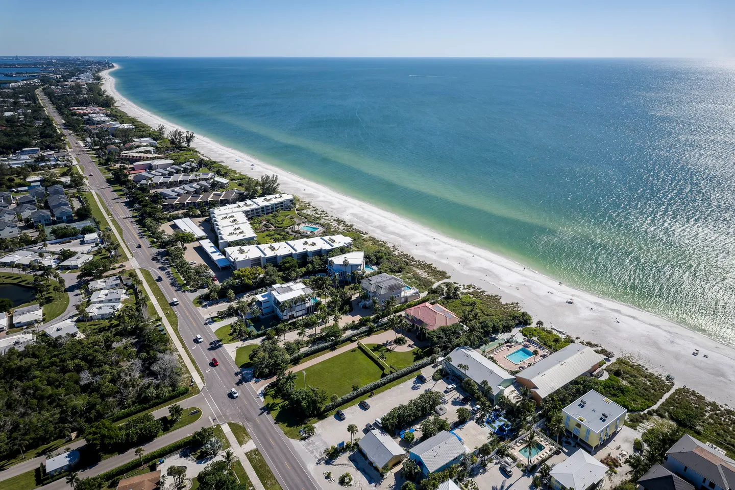 Aerial view of a coastal road, residential properties, sandy beach, and calm ocean water under a clear blue sky.