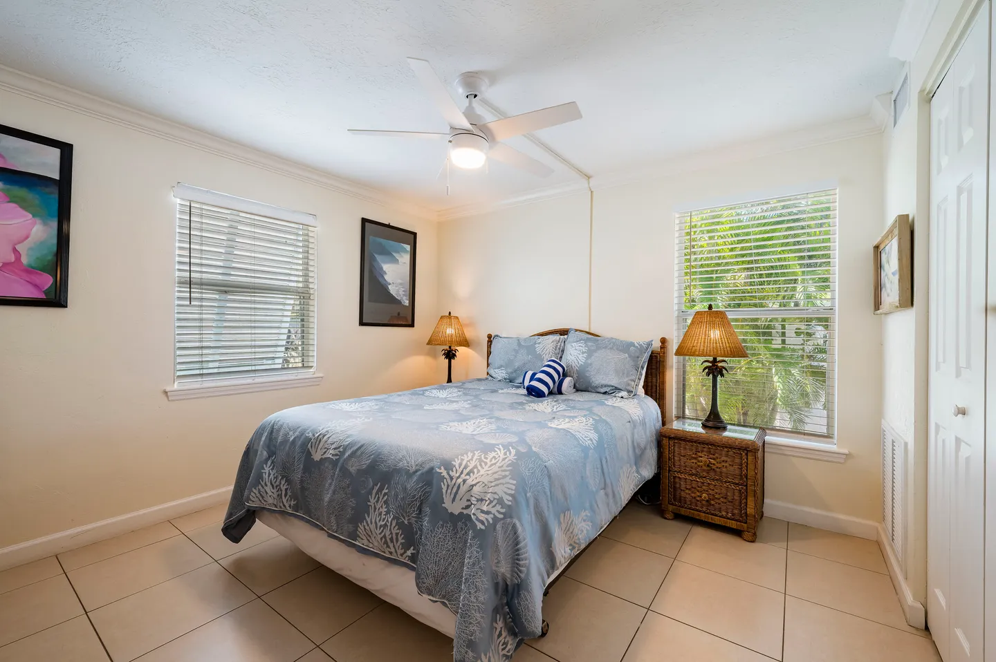 Bedroom with tile flooring, a bed dressed in a blue coastal-themed spread, two windows with blinds, and a white ceiling fan.