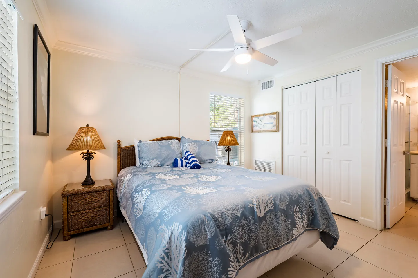 Bedroom with tile flooring, a bed made with a blue coastal-themed comforter, two nightstands with lamps, a ceiling fan, and white closet doors.