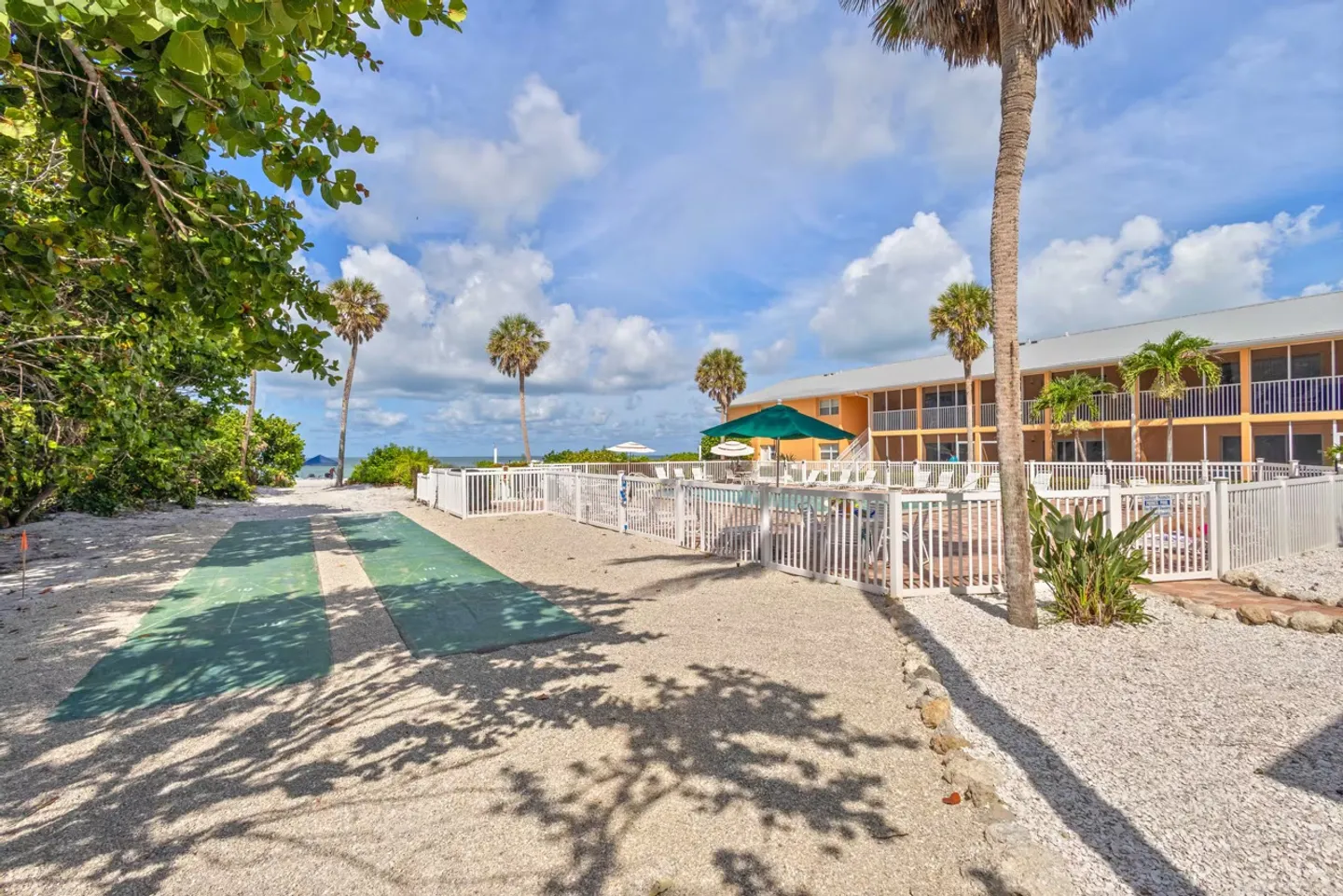 Outdoor recreation area with green shuffleboard courts, fenced swimming pool, palm trees, and residential building under a blue sky.
