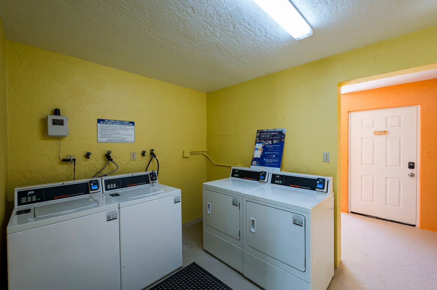 A laundry room with yellow walls, two commercial washing machines, and two commercial dryers.