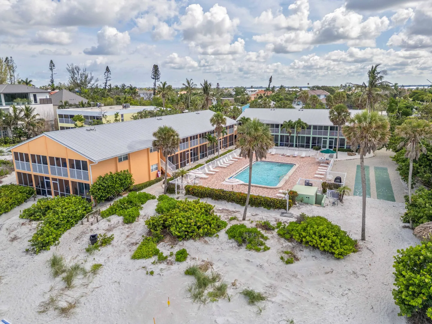 Aerial view of a coastal multi-unit residential complex featuring a swimming pool, patio, and sandy grounds with palm trees.