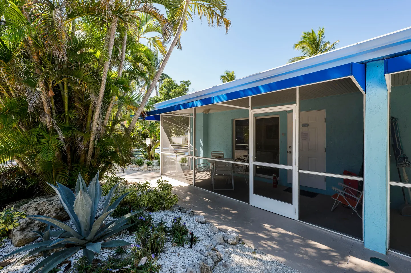 Exterior view of a light blue building with a screened-in porch, patio furniture, and surrounding tropical landscaping.