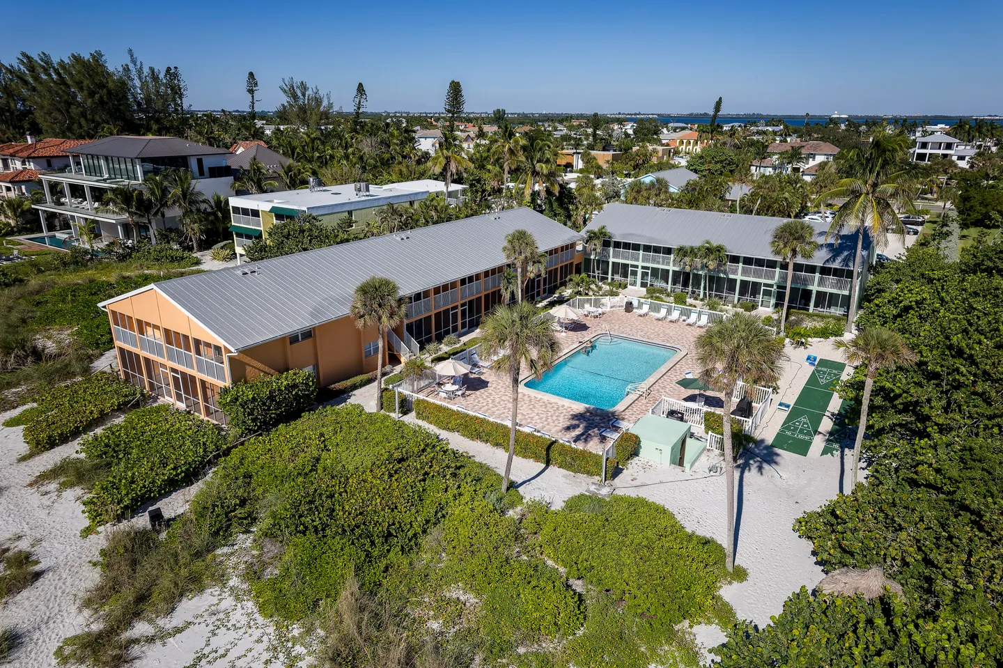 Aerial view of a coastal property featuring two two-story apartment buildings, a swimming pool surrounded by a patio, shuffleboard courts, and sandy grounds with palm trees.