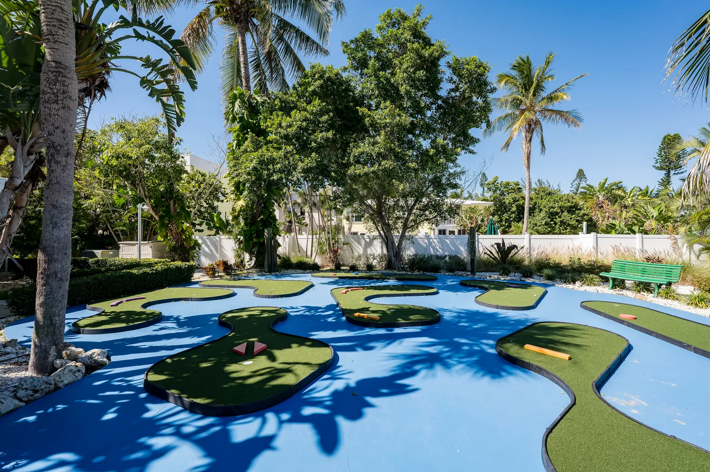 Outdoor miniature golf course with green artificial turf putting areas surrounded by a blue surface and tropical trees.