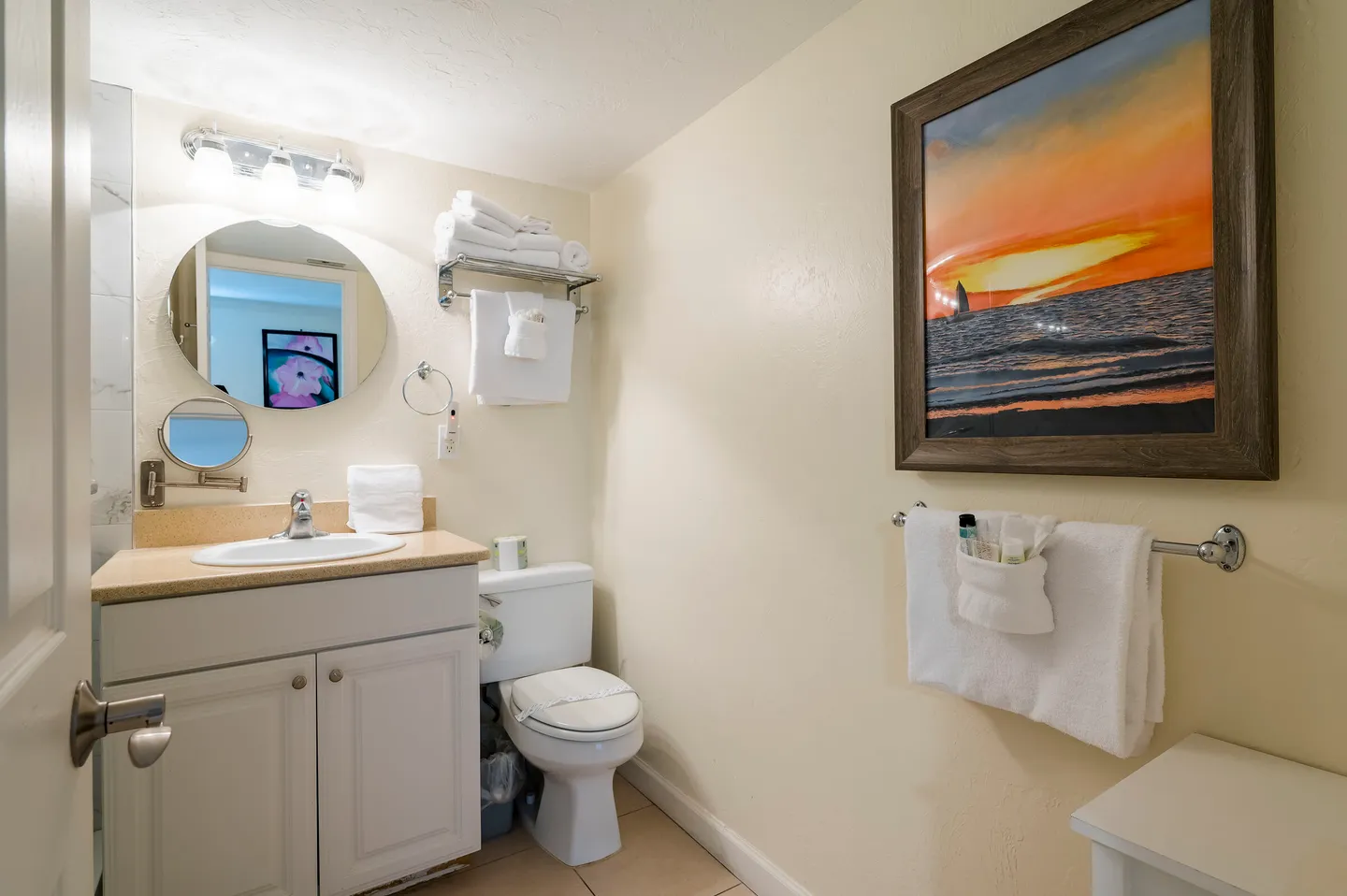Bathroom with a white vanity, round mirror, toilet, and a framed sunset painting on the wall.