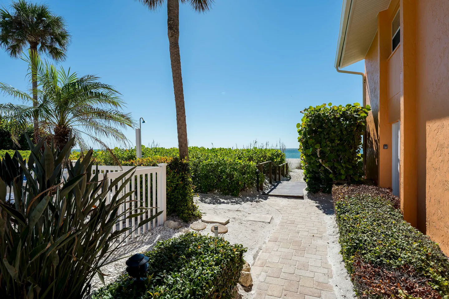A stone paver path leads past green foliage, a palm tree, and a small wooden bridge toward the sandy beach and ocean under a clear blue sky.