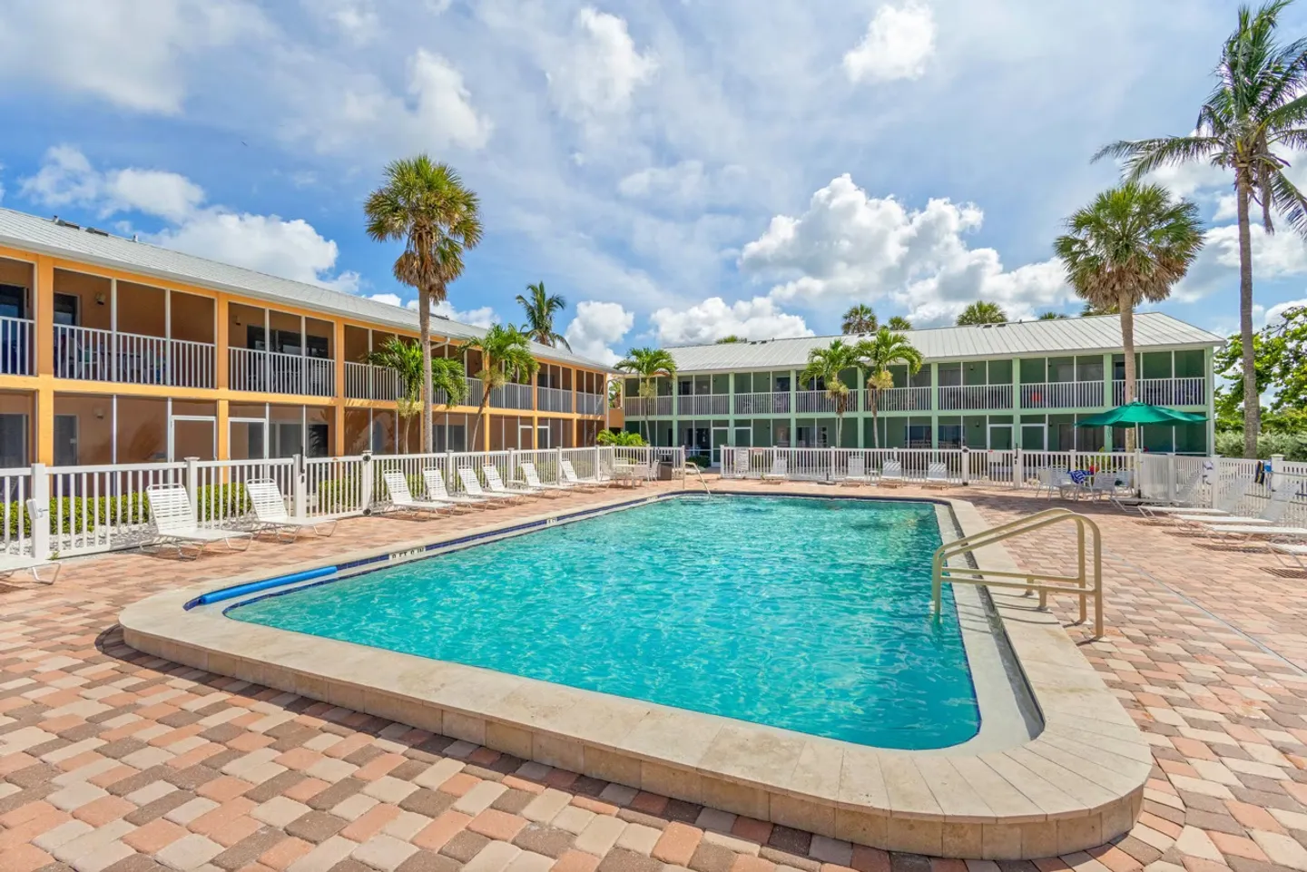 Outdoor swimming pool surrounded by a paver patio and two-story apartment buildings under a partly cloudy sky.