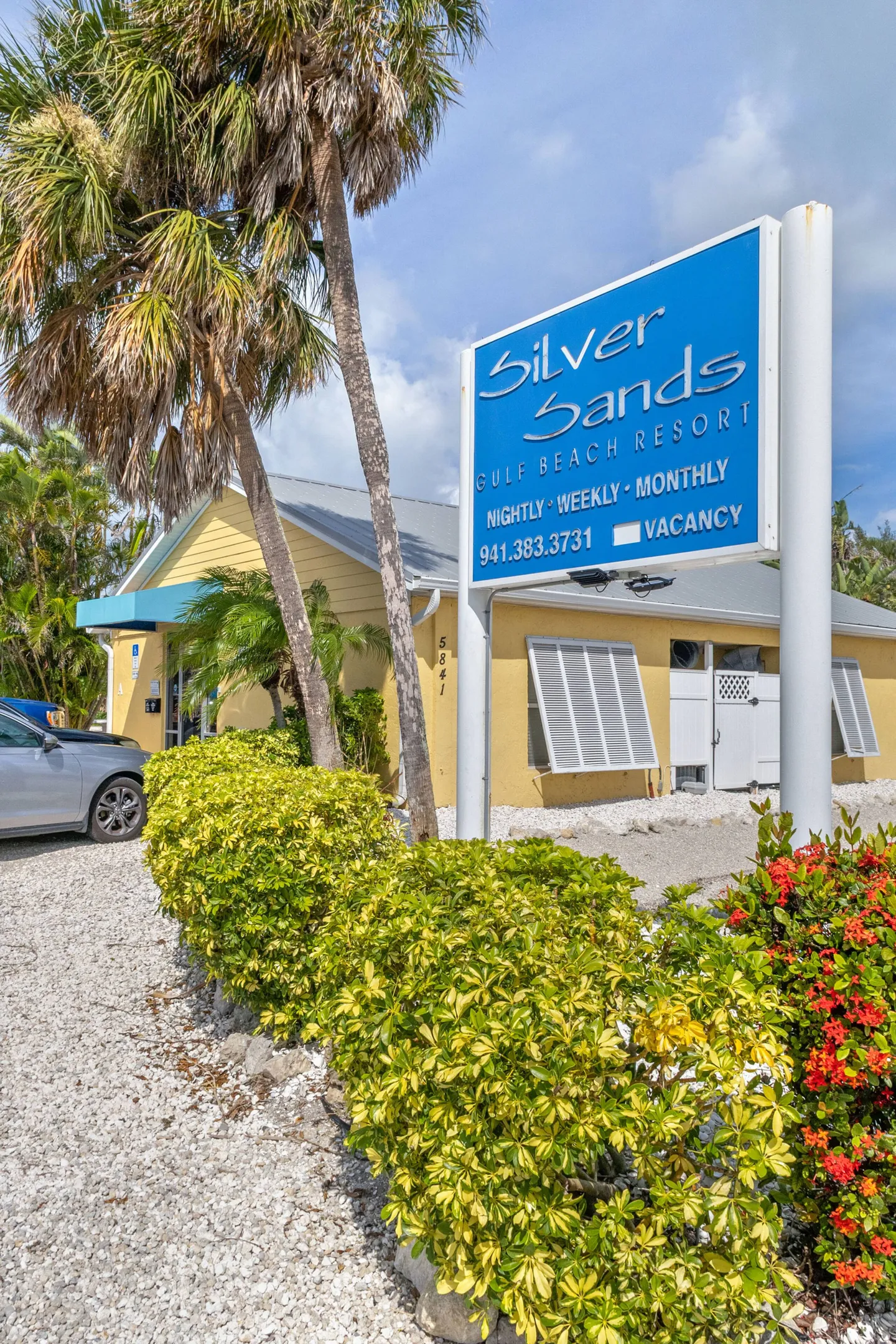 Property entrance sign for Silver Sands Gulf Beach Resort next to tropical landscaping and a yellow building.