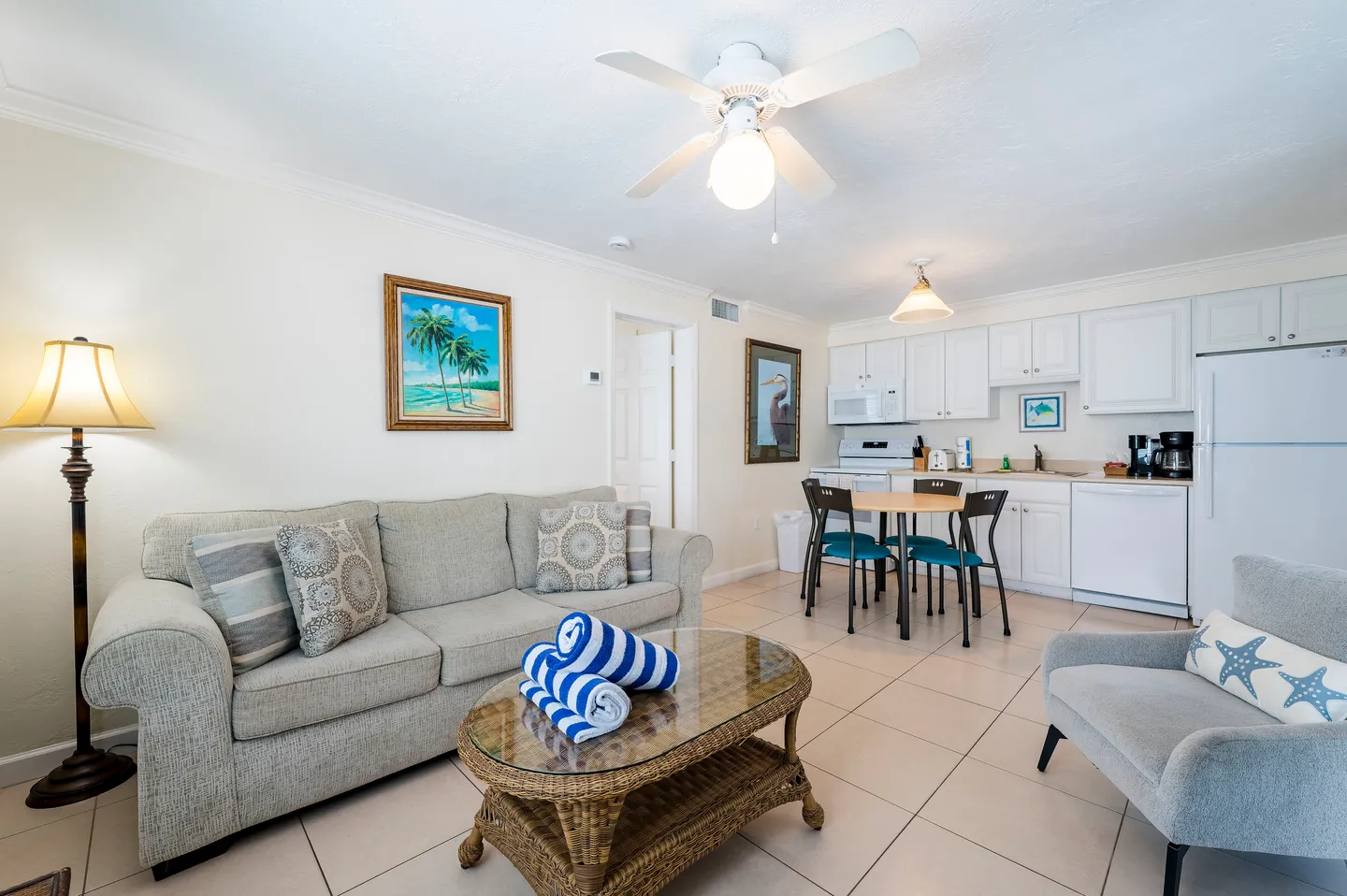 Open-concept living room with a sofa, coffee table, armchair, small dining table, and adjacent kitchen area with white cabinets.