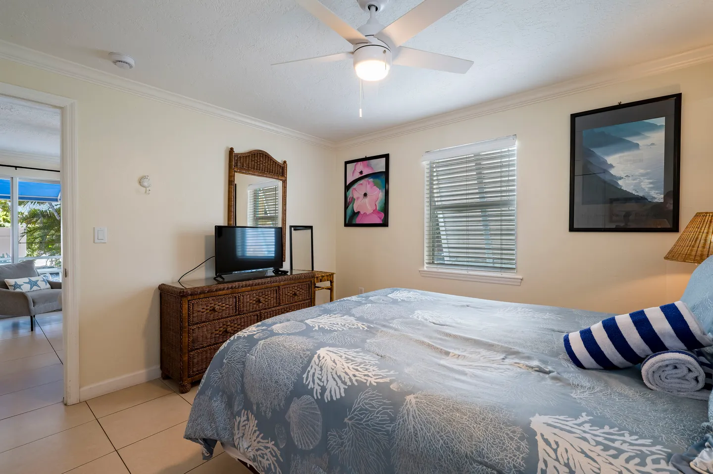 Bedroom with tile flooring, a bed with coastal-patterned bedding, a wicker dresser with a TV, and a white ceiling fan.