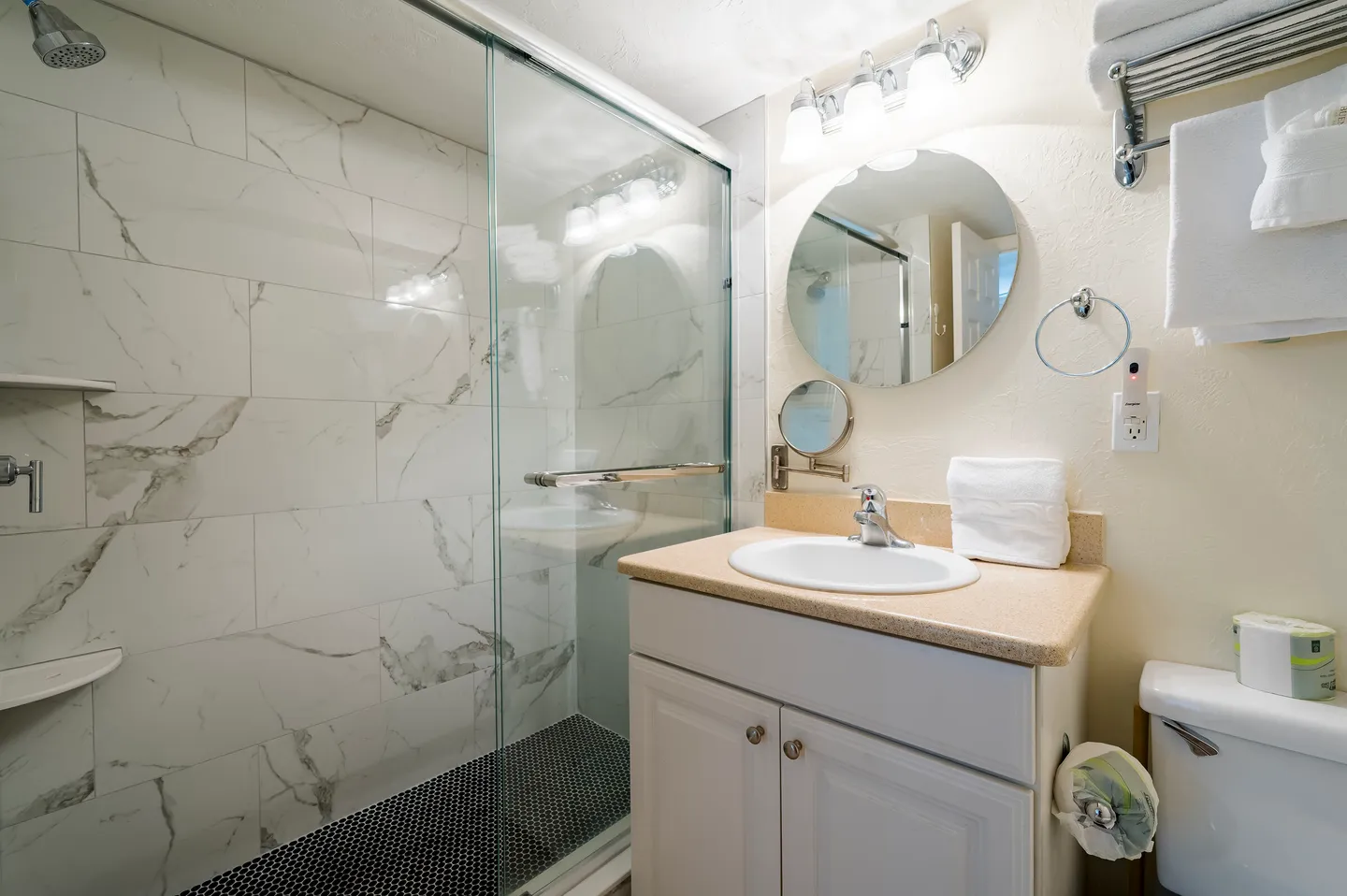 Bathroom with a glass-enclosed walk-in shower featuring marble-style wall tiles and a single-sink vanity with a round mirror.