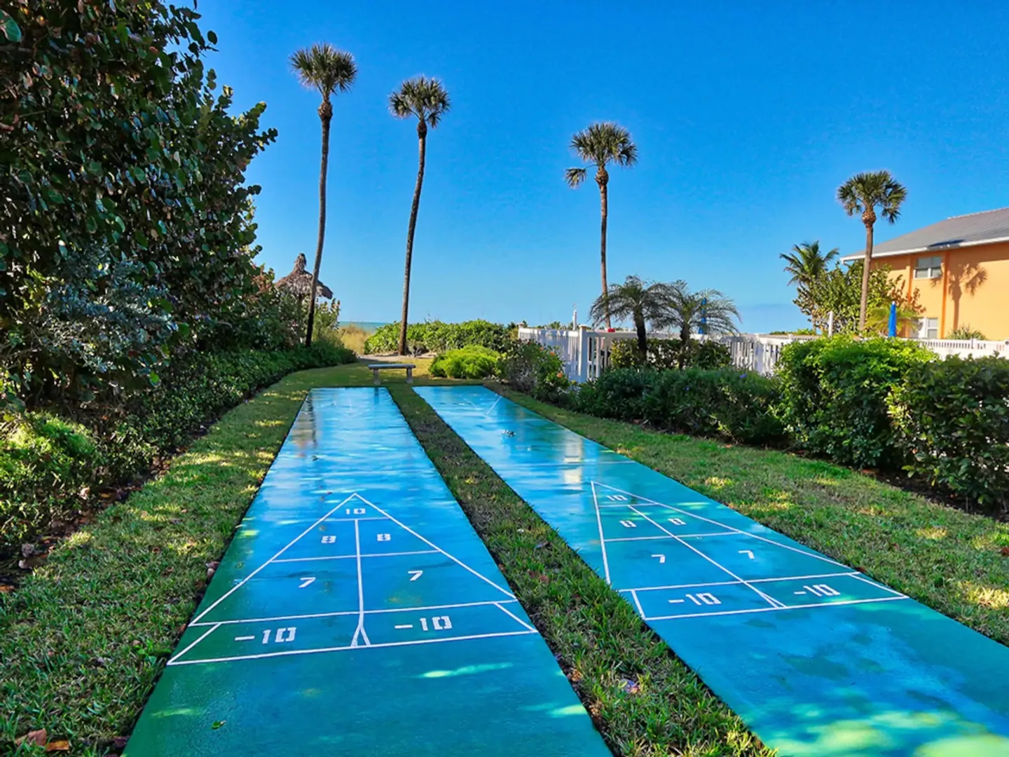 Two outdoor shuffleboard courts surrounded by grass, palm trees, and tropical landscaping under a clear blue sky.