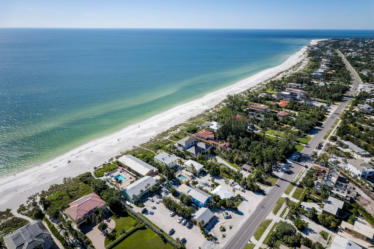 High aerial view of a white sand coastline with turquoise ocean water, beach houses, palm trees, and a road.