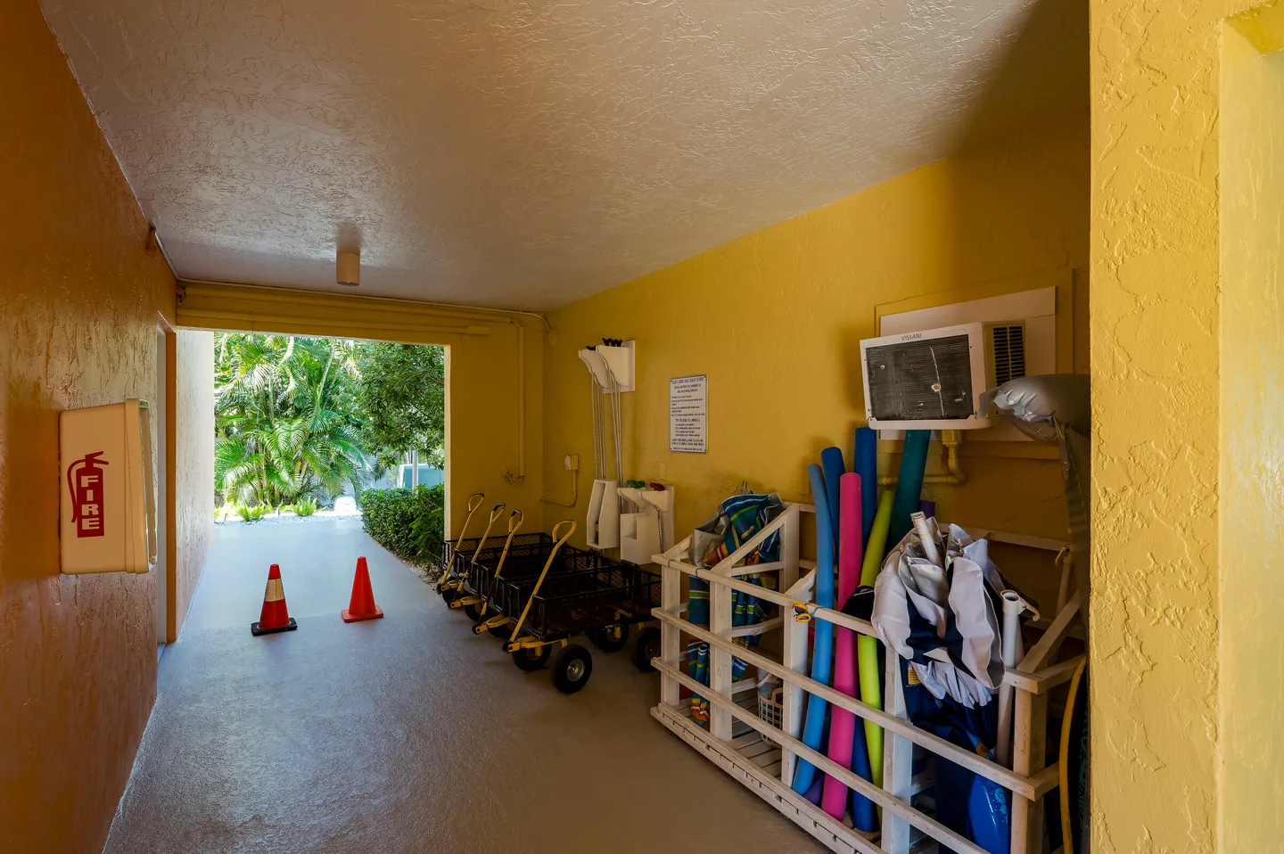A covered breezeway with yellow walls storing rolling carts, pool noodles, and beach gear leading out to a tropical garden.