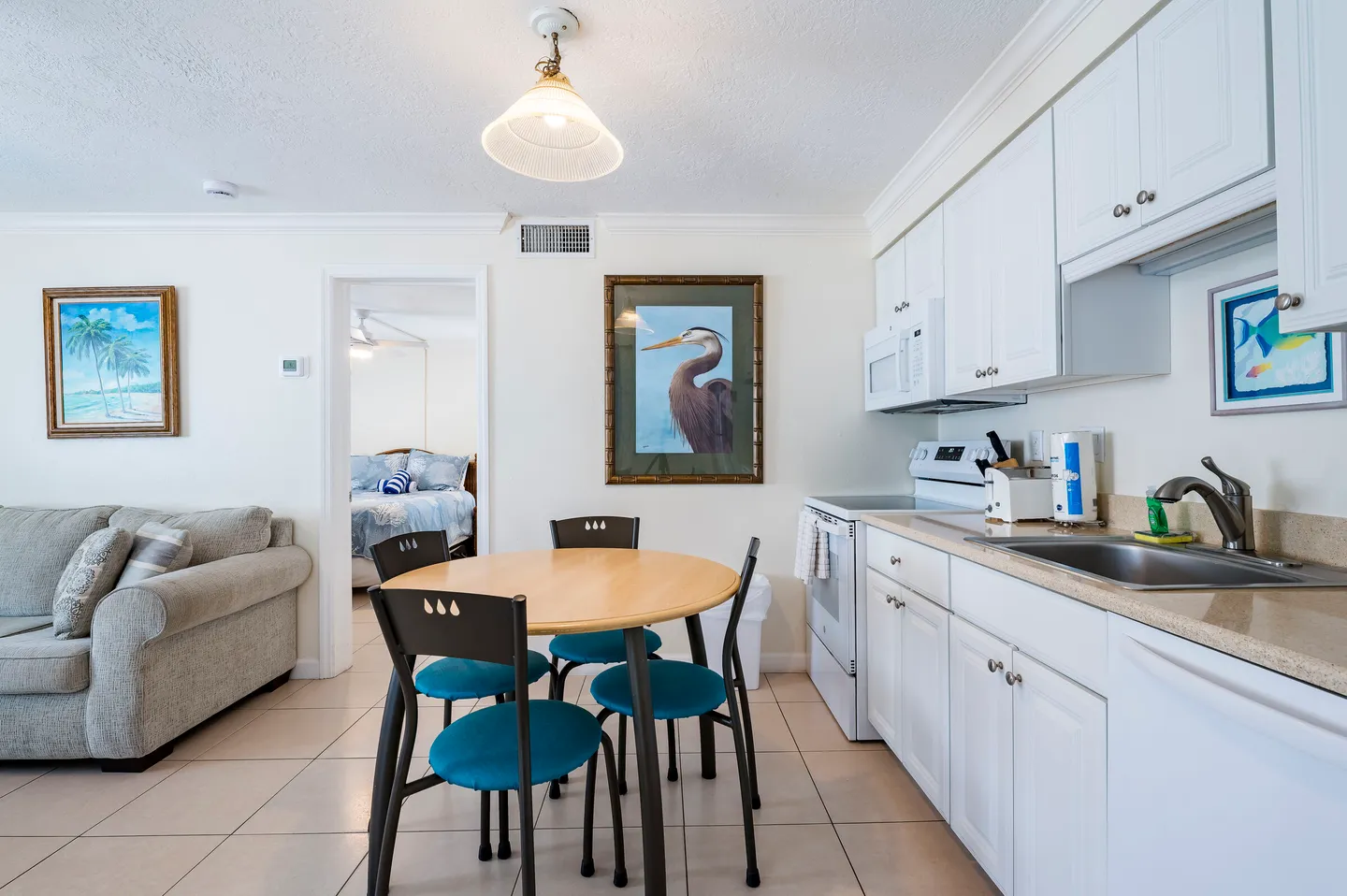 Dining area with round wooden table and turquoise-cushioned chairs adjacent to kitchen with white cabinets, near living room and open doorway to bedroom.