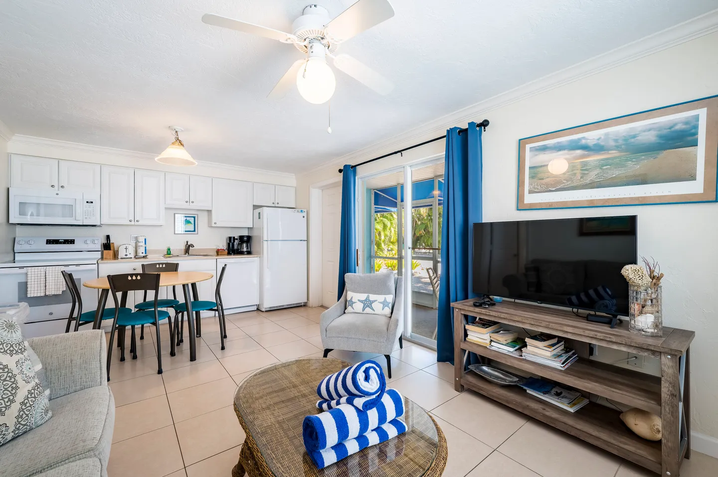 Open living, dining, and kitchen area with tiled floors, a dining table, armchair, TV console, and sliding glass doors with blue curtains.
