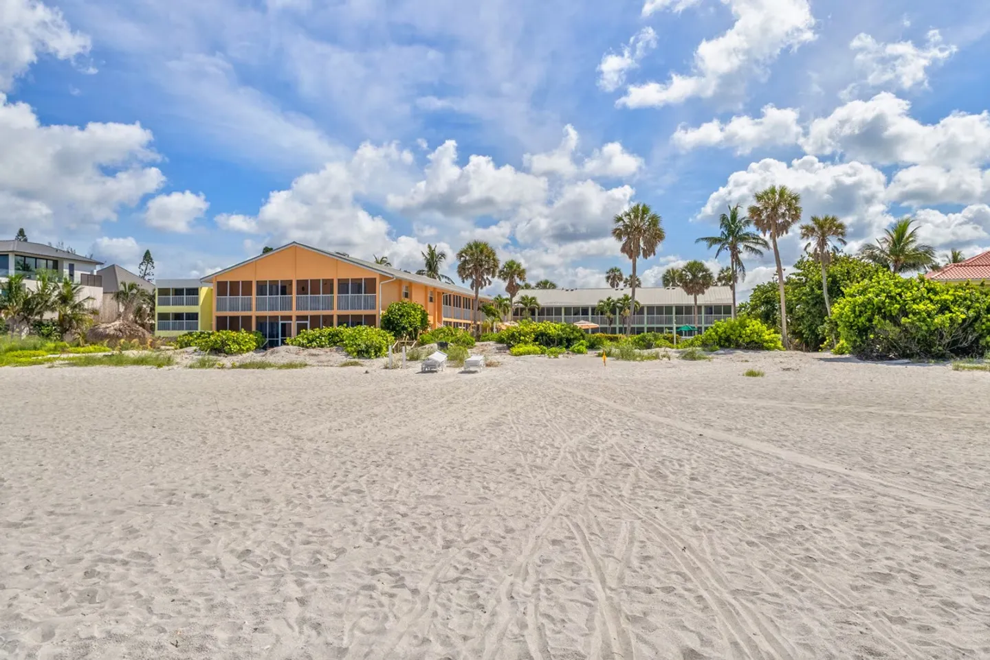 Exterior view of beachfront residential buildings with palm trees and a sandy beach under a blue sky.