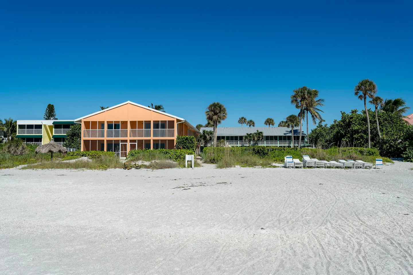 Exterior view of beachfront residential buildings with balconies and palm trees situated directly on the white sand beach under a clear blue sky.