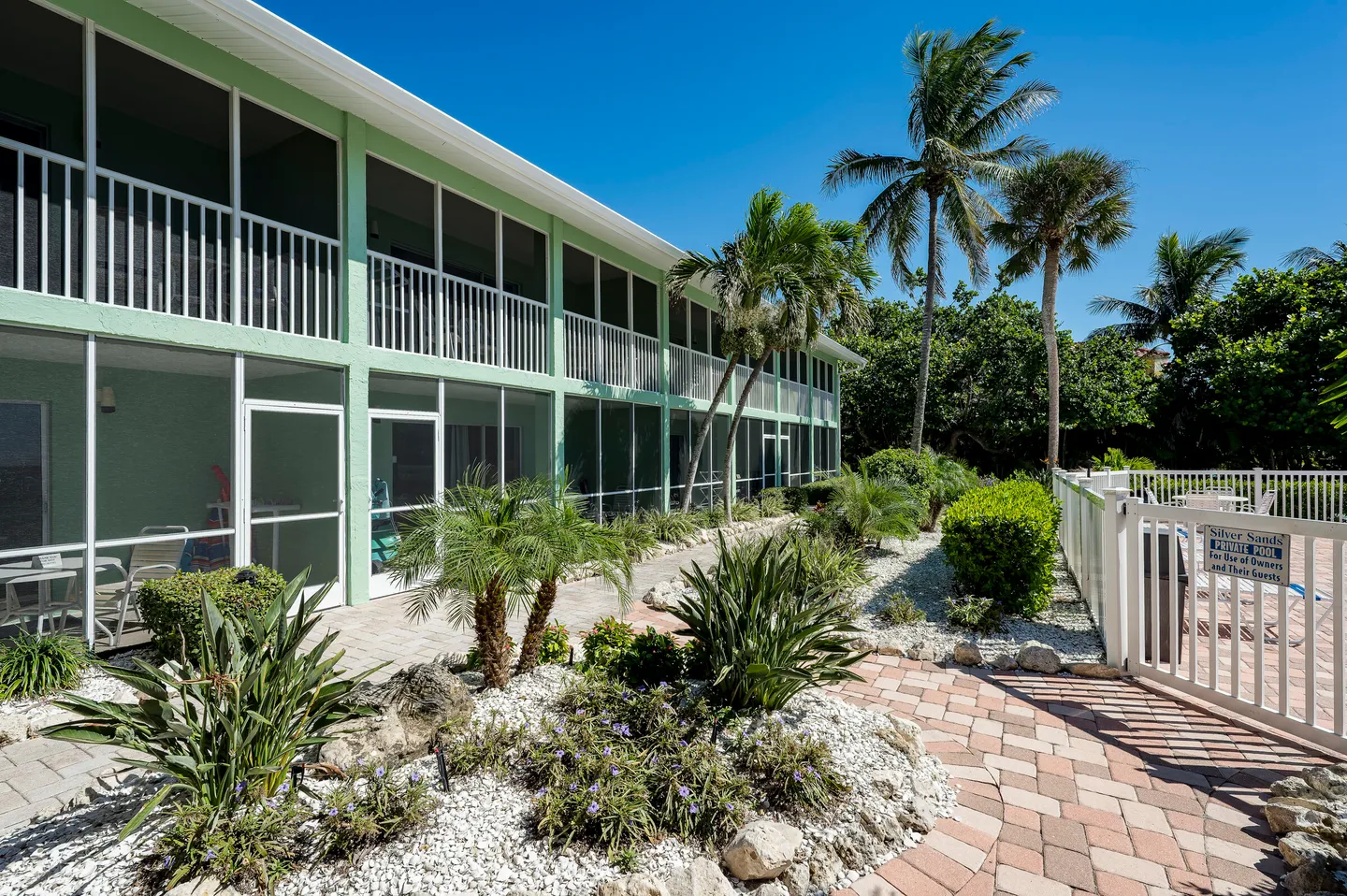 Exterior view of a light green two-story building with screened porches, landscaped path, and a fenced pool area.
