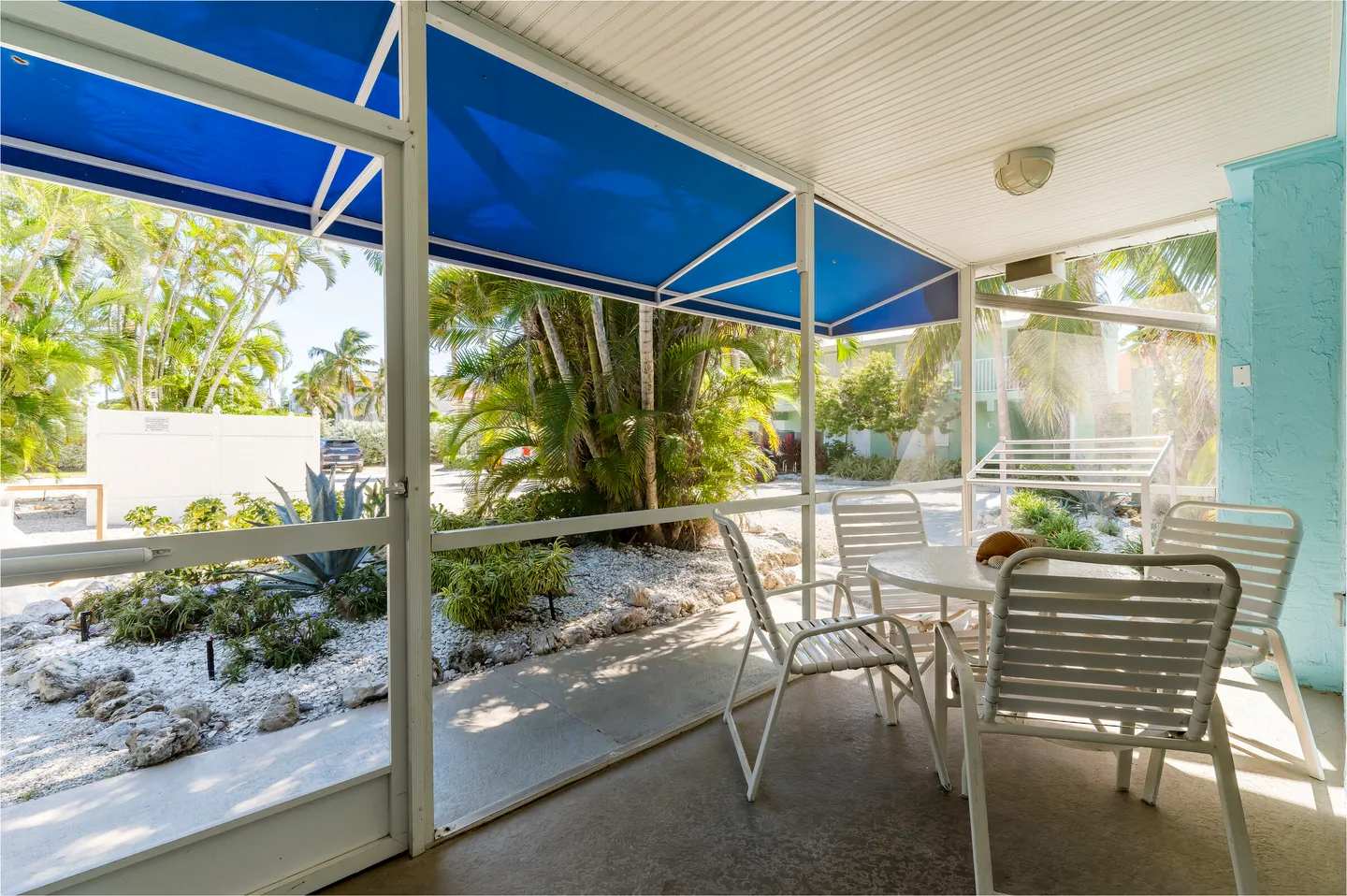 Screened porch furnished with a white round patio table and matching chairs, overlooking tropical landscaping.
