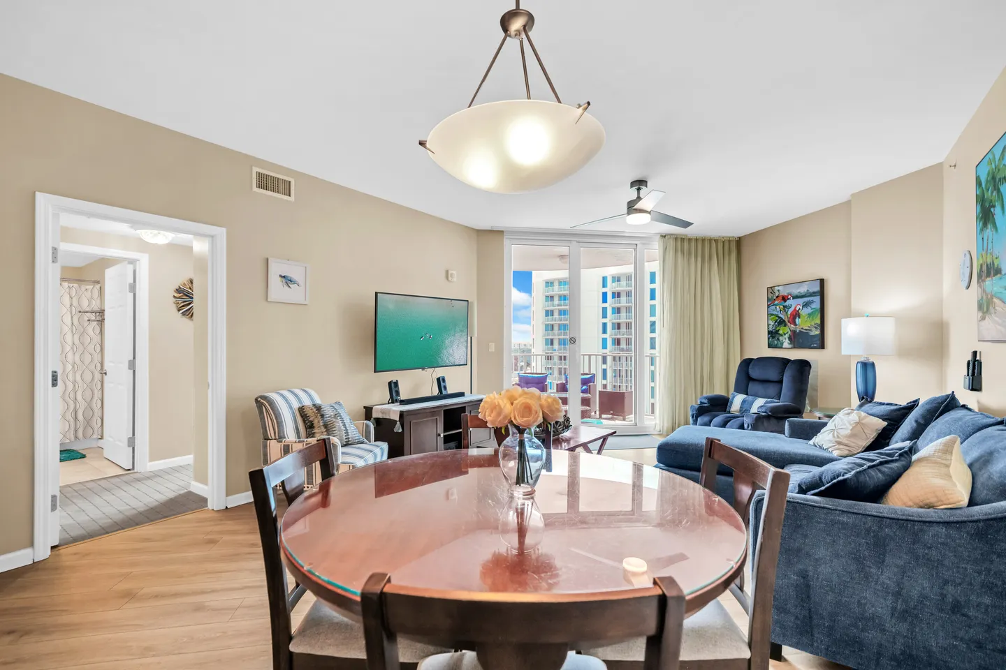 Dining area with a round wooden table in the foreground overlooking an open-plan living room with a blue sectional sofa and balcony doors.